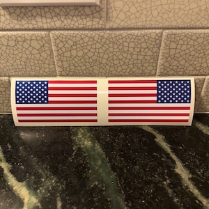 May include: Two American flag stickers on a white background. The stickers are curved to fit a rounded surface.