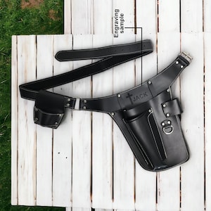 Op de afbeelding: Zwarte leren gereedschapsriem met een klein vak en een groter vak met een metalen ring. De riem heeft witte stiksels en is gegraveerd met de naam "JACK" en een kruis.