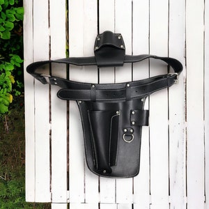Könnte beinhalten: Schwarzer Leder-Werkzeuggürtel mit weißen Nähten. Der Gürtel verfügt über mehrere Taschen und Schlaufen für Werkzeuge, darunter eine Halterung für ein kleines Werkzeug und eine größere Tasche. Eine Metallschnalle sichert den Gürtel.