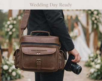 May include: A brown leather camera bag with a top handle and shoulder strap, featuring a front flap pocket with a buckle closure and the text ADAMS PHOTOGRAPHY embossed on the front. The bag has side pockets and black zipper accents.