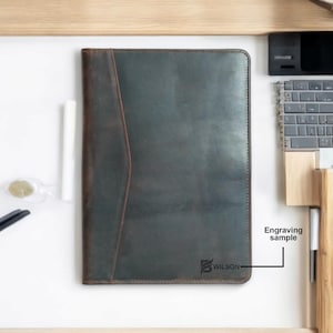 May include: A dark green leather laptop sleeve with a brown leather flap and a pocket on the front. The sleeve has a stitched edge and the text "WILSON" is engraved on the bottom right corner. Engraving sample.