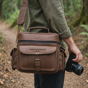 May include: A brown leather camera bag with a top handle and shoulder strap, featuring a front pocket with a buckle closure and the text ADAMS PHOTOGRAPHY embossed on the flap. The bag includes side pockets and a black zipper closure.