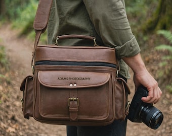 May include: A brown leather camera bag with a top handle and shoulder strap, featuring a front pocket with a buckle closure and the text ADAMS PHOTOGRAPHY embossed on the flap. The bag includes side pockets and a black zipper closure.