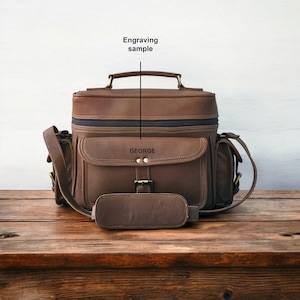 Könnte beinhalten: Braune Leder-Umhängetasche mit oberem Griff, Schulterriemen und mehreren Taschen. Die Tasche hat eine Frontklappe mit Schnallenverschluss und der Name "GEORGE" ist auf der Vorderseite eingraviert. Der Text "Engraving sample" steht über der Tasche.