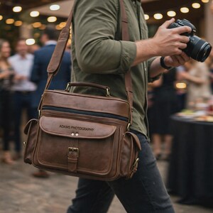 Puede incluir: Una bolsa de cuero marrón para cámara con correa ajustable, asa superior y bolsillos laterales. La solapa frontal tiene un cierre de hebilla y el texto ADAMS PHOTOGRAPHY grabado en negro. La bolsa tiene una forma rectangular.