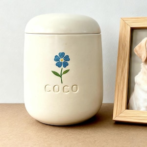 Puede incluir: Urna para mascotas de cerámica blanquecina con un diseño floral azul y el nombre "COCO" en relieve. Junto a la urna, un marco de madera con una foto de un Labrador Retriever.