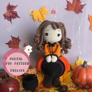 Könnte beinhalten: Häkelanleitung für eine Hexenpuppe mit braunen Haaren, die ein orange-schwarzes Kleid und einen schwarzen Hexenhut trägt. Die Puppe sitzt auf einem Bett aus Herbstlaub mit einem Kürbis und einem Kessel.