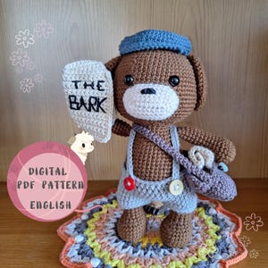 Könnte beinhalten: Häkelanleitung für einen braunen Bären mit blauem Hut und Latzhose, der ein Schild mit der Aufschrift "The Bark" hält. Der Bär steht auf einem bunten Häkelteppich.