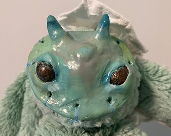 Könnte beinhalten: Ein hellblaues Plüschtier mit einem modelliertem Kopf, der an einen Frosch erinnert. Der Kopf hat große, braune Augen und einen genähten Mund. Das Spielzeug trägt einen weißen Stoffhut.