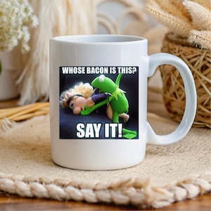 Peut inclure: Mug en céramique blanche avec une image humoristique de Kermit la Grenouille et Miss Piggy, avec le texte "WHOSE BACON IS THIS? SAY IT!". La tasse a une grande anse et est conçue pour les boissons chaudes.