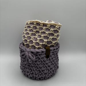 Op de afbeelding: Twee handgemaakte gehaakte manden. De onderste mand is effen grijs met een decoratieve knoop en een bruin leren label. De bovenste mand is lichtpaars met een crèmekleurig, open geweven ontwerp. Beide manden zijn rond.
