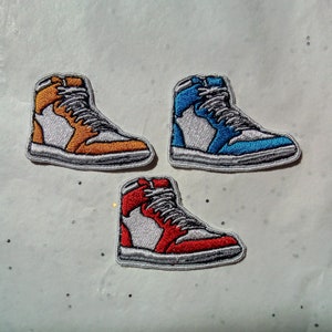 May include: Three embroidered patches of high-top trainers in orange, blue, and red. Each patch features a white and grey design with a black outline. The patches are on a white surface.
