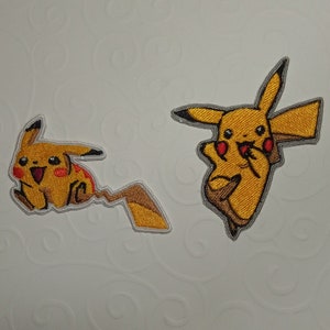 Peut inclure: Deux écussons Pikachu brodés. L'un montre Pikachu courant avec une expression joyeuse, l'autre montre Pikachu avec un doigt dans la bouche. Les deux écussons sont jaunes avec des joues rouges, des queues marron et des contours gris.