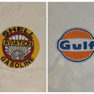 Puede incluir: Dos parches bordados. Uno es redondo con el texto "SHELL AVIATION GASOLINE", un borde amarillo y un diseño rojo y blanco. El otro tiene forma ovalada con "GULF" en texto azul, un centro naranja y un borde azul.