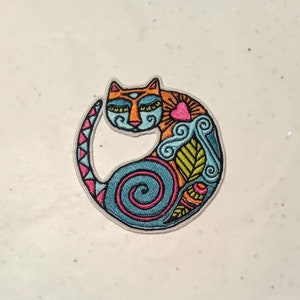 Puede incluir: Parche bordado con un gato estilizado en posición circular. El gato tiene la cabeza naranja, el cuerpo azul y un corazón rosa. El diseño incluye espirales, hojas y una cola rosa y azul. El parche está sobre un fondo blanco.