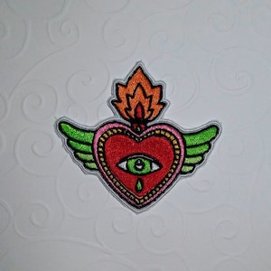Puede incluir: Parche bordado con un corazón rojo con un ojo verde, alas verdes y llamas naranjas. El corazón tiene un borde plateado y un contorno rosa. El parche está sobre un fondo blanco con un patrón de remolino.