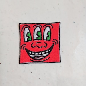 Könnte beinhalten: Ein rotes quadratisches Patch mit einem Cartoon-Gesicht. Es hat drei grüne Augen, eine Nase und ein breites, zahnreiches Grinsen. Der Patch ist mit schwarzen und weißen Details bestickt und schafft ein verspieltes Design. Geeignet für Kleidung oder Accessoires.