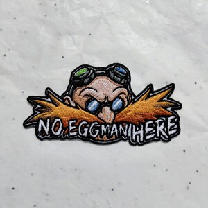 May include: An embroidered patch featuring a cartoon character with a large orange moustache, round blue glasses, and black goggles on their forehead. The text NO EGGMAN HERE is stitched in white capital letters across the bottom of the design.