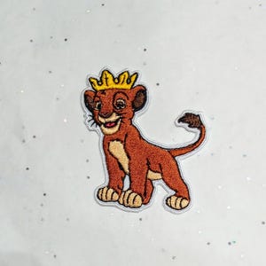 Peut inclure: Écusson brodé représentant un lionceau de dessin animé portant une couronne dorée. Le lionceau est brun avec un ventre beige et une expression souriante. L'écusson a une bordure blanche et est posé sur un fond blanc.