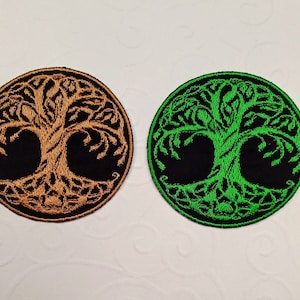 Può includere: Due toppe rotonde ricamate con un disegno ad albero. Una toppa presenta un albero dorato su sfondo nero, l'altra un albero verde su nero. Le toppe hanno un aspetto dettagliato e strutturato.