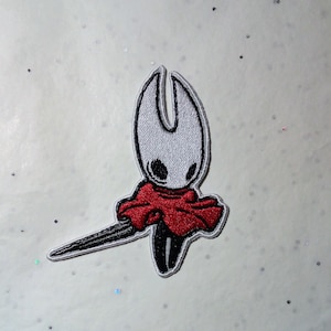 Peut inclure: Écusson brodé représentant un personnage stylisé avec une tête blanche, des yeux noirs et une cape rouge. Le personnage tient une épée noire et grise. L'écusson a une bordure blanche et est destiné aux vêtements ou accessoires.
