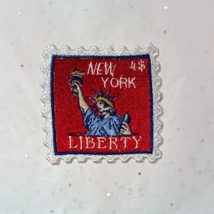 May include: Embroidered patch in the shape of a postage stamp. The patch features a red background with the words "NEW YORK" and "LIBERTY" in white. The image of the Statue of Liberty is in the center, with a blue robe and a golden crown.