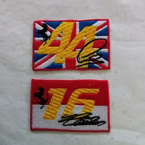 Peut inclure: Deux écussons rectangulaires brodés. L'écusson supérieur présente un fond Union Jack avec un numéro 44 jaune, un logo de cheval cabré noir et une signature noire. L'écusson inférieur a un fond rouge et blanc, un numéro 16 jaune, un logo de cheval cabré noir et une signature noire.