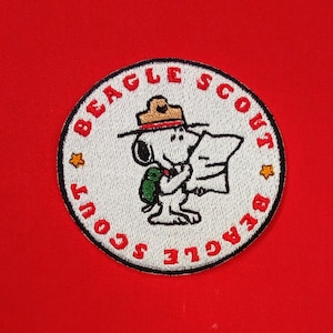 May include: Embroidered patch featuring Snoopy in a scout uniform, holding a map. The patch is circular with the words "BEAGLE SCOUT" in red around the edge. Snoopy is wearing a brown hat and a green backpack. Two gold stars are on the patch.