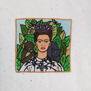 Peut inclure: Écusson brodé représentant une femme avec un singe et un chat noir. La femme a les cheveux foncés et une couronne florale. Le fond comprend des feuilles vertes et un ciel bleu clair. L'écusson a une bordure marron.