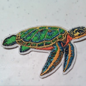Può includere: Toppa ricamata a forma di tartaruga marina con bordo bianco. La tartaruga presenta un guscio verde e blu, pinne arancioni e blu e una testa gialla. Questa toppa decorativa può essere stirata o cucita su vestiti o accessori.