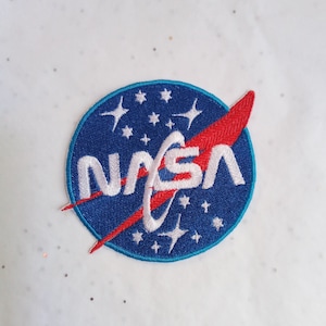 Pode incluir: Patch da NASA bordado com o logotipo da NASA em branco, vermelho e azul. O patch é circular com fundo azul, estrelas brancas e o texto NASA em branco. Um swoosh vermelho cruza o logotipo.