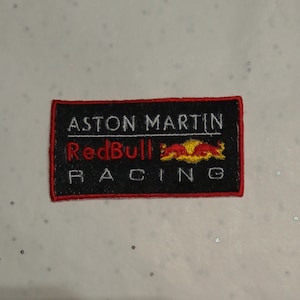 Puede incluir: Parche bordado con el logotipo de Aston Martin Red Bull Racing. El parche rectangular tiene un fondo negro con texto blanco que dice "ASTON MARTIN" y "RACING". La palabra "Red Bull" es roja y el logotipo de Red Bull es amarillo y naranja.