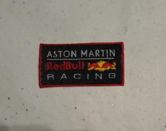 May include: Embroidered patch featuring the Aston Martin Red Bull Racing logo. The rectangular patch has a black background with white text that reads "ASTON MARTIN" and "RACING". The word "Red Bull" is in red, and the Red Bull logo is in yellow and orange.