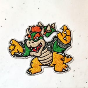 May include: Embroidered patch featuring Bowser, a character from the Super Mario video game series. The patch depicts Bowser in a dynamic pose, with a green and orange color scheme, and a detailed design. The patch is outlined with a white border.