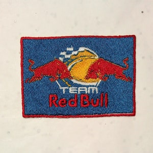 May include: Embroidered patch featuring the Red Bull logo. The patch has a blue background with a red border. Two red bulls face each other above a yellow and white design. The words "TEAM Red Bull" are embroidered in white.