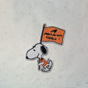 May include: Embroidered patch featuring Snoopy holding a flag with the McLaren Formula 1 logo. The patch has a white background, a black and white Snoopy, and an orange flag with black text.