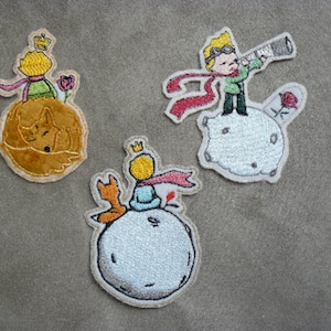 Peut inclure: Trois écussons brodés représentant des personnages du "Petit Prince". L'un montre le prince avec un renard, un autre avec un télescope, et le troisième avec une rose et un renard endormi. Chaque écusson a une bordure blanche sur fond gris.