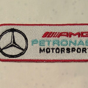 Può includere: Toppa ricamata con il logo Mercedes-Benz e la scritta "AMG PETRONAS MOTORSPORT". La toppa è rettangolare con bordo rosso e sfondo grigio chiaro. Il logo è nero e il testo è rosso e verde acqua.