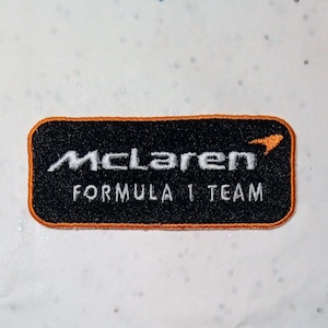 Op de afbeelding: Geborduurde patch met het McLaren Formula 1 Team logo. De patch heeft een zwarte achtergrond, witte tekst en een oranje rand. Het McLaren-logo en de woorden "FORMULA 1 TEAM" worden weergegeven.