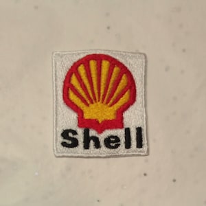 May include: Embroidered patch featuring the Shell logo. The logo is a red and yellow scallop shell design on a white background. The word "Shell" is embroidered in black letters below the shell.
