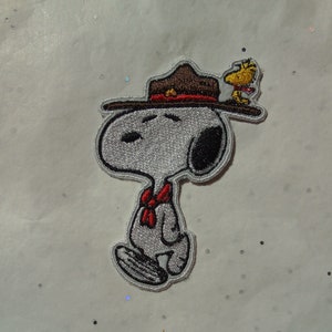 Può includere: Toppa ricamata con Snoopy con un cappello marrone con una fascia rossa e Woodstock appollaiato sopra. Snoopy indossa una sciarpa rossa. La toppa ha uno sfondo bianco con contorni neri.