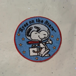 Puede incluir: Parche bordado circular con Snoopy con casco de astronauta y bufanda roja, sosteniendo un maletín. El parche azul tiene un borde rojo y el texto "Eyes on the Stars".