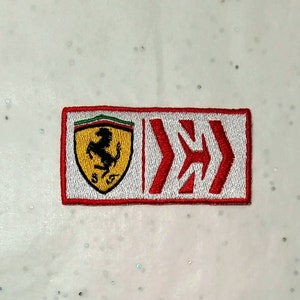 Op de afbeelding: Geborduurde patch met het Ferrari-logo, een zwart steigerend paard op een gele schild met een rode rand. De patch bevat ook een sectie met rode pijlen op een witte achtergrond en een rode rand.