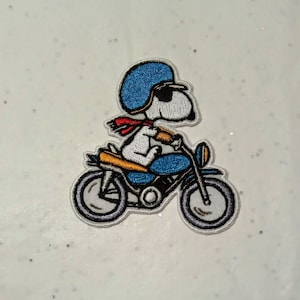Könnte beinhalten: Gestickter Aufnäher mit Snoopy aus Peanuts auf einem blauen Motorrad. Snoopy trägt einen blauen Helm, eine Sonnenbrille und einen roten Schal. Das Motorrad hat einen blauen Rahmen mit gelben und schwarzen Details und grauen Rädern.