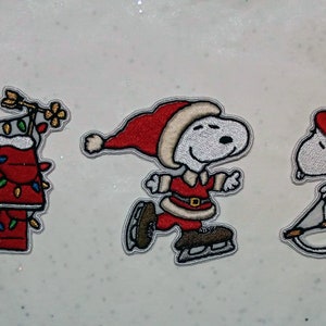 Op de afbeelding: Geborduurde patches met Snoopy in feestelijke kerstscènes. Eén patch toont Snoopy met zijn hondenhok versierd met lichtjes, een andere toont Snoopy schaatsend en de laatste toont Snoopy aan het sleeën. Elke patch heeft een witte rand.