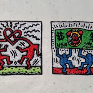 May include: Two embroidered patches with Keith Haring-style figures. One features two red figures forming a heart, the other shows blue figures holding up a green dollar bill with a cartoon character and the text "USA".