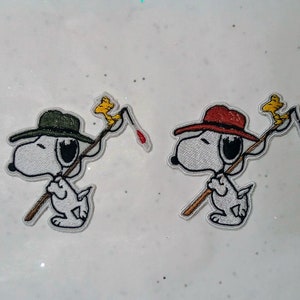 Peut inclure: Écussons brodés représentant Snoopy, le beagle de dessin animé, en pêcheur. L'un porte un chapeau vert et un poisson rouge, l'autre un chapeau rouge et un poisson bleu. Les deux écussons ont un oiseau jaune sur la canne à pêche.