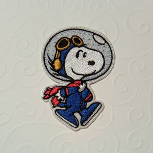 Puede incluir: Parche bordado con Snoopy con un traje de piloto azul, bufanda roja y casco con gafas. El parche tiene un fondo blanco y está contorneado con hilo plateado. El personaje de dibujos animados está sonriendo y caminando.
