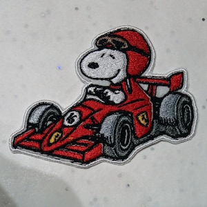 Peut inclure: Écusson brodé représentant Snoopy dans une voiture de course rouge avec le numéro 16. Snoopy porte un casque et des lunettes rouges. La voiture porte le logo Ferrari. L'écusson a une bordure blanche et est sur fond blanc.