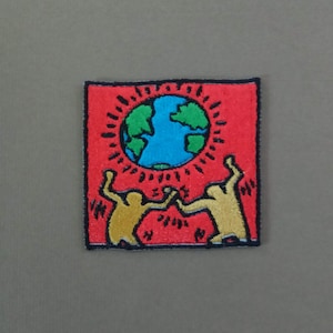 Op de afbeelding: Een vierkante geborduurde patch met een felrode achtergrond en een centrale illustratie van een blauwe en groene wereldbol. Twee gestileerde beige figuren met opgeheven armen staan onder de wereldbol, omringd door zwarte stralende lijnen.
